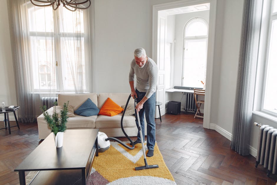 A yellow handheld vacuum cleaner with a hose and attachment is placed on a beige floral area rug in a living room, near a wooden media console with black metal legs. The console holds a remote control
